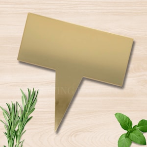 May include: A gold-coloured plant marker with a rectangular top and a pointed base. The marker is angled slightly, casting a shadow. Rosemary and basil sprigs are visible, suggesting a garden or culinary theme.