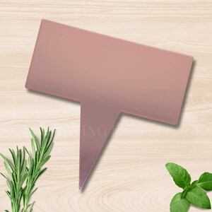 May include: A pink rectangular garden stake with a pointed bottom, suitable for plant labels. The word "ING" is partially visible on the stake. Rosemary and basil sprigs are either side of the stake, set against a light wood grain background.