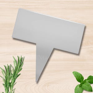 May include: A silver metal plant marker with a rectangular top and a pointed bottom. The marker is blank, ready for labeling. Rosemary and basil sprigs are visible in the background, suggesting a garden theme.