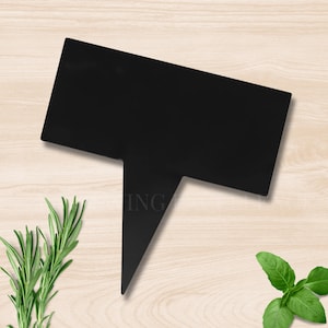 May include: A black rectangular garden marker with a pointed stake. The word "ING" is partially visible on the marker. Fresh rosemary and basil sprigs are on a light wood surface.