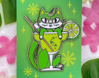 May include: Green enamel pin of a cat wearing a sombrero and sitting in a margarita glass with a lime wedge. The pin says "Kitty Cocktails Enamel Pin MEOWGARITA" and "www.pinkowlet.com".