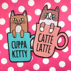 Op de afbeelding: Twee geborduurde strijkpatches met katten in mokken. Eén patch is turquoise blauw met een bruine kat en de tekst "Cuppa Kitty". De andere patch is perzikkleurig met een bruine en witte kat en de tekst "Catte Latte".