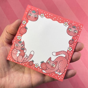 May include: A square notepad with a red border featuring white polka dots and a scalloped edge. The notepad is decorated with illustrations of pink cats and strawberries. The cats are holding strawberries, and small white flowers are also included in the design.
