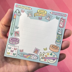 May include: A light blue notepad with a cat-themed design. The notepad features illustrations of cat stickers, stamps, pens, tape, and glue. The notepad is held in a person's hand.
