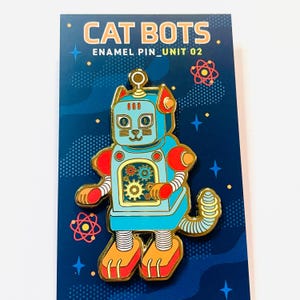 May include: Enamel pin featuring a blue robot cat with red and orange accents. The pin has a gold outline and intricate gear details on its chest. The pin is on a dark blue card with the words "CAT BOTS" and "ENAMEL PIN_UNIT 02".
