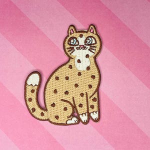 May include: Embroidered patch of a seated cat with a tan body and brown spots. The cat has a white chest and paws, with blue eyes and brown whiskers. The patch is outlined in brown and set against a pink and white striped background.