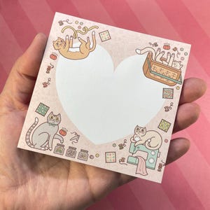 May include: A pink heart-shaped notepad featuring cartoon cats and sewing-themed illustrations. The notepad has a blank heart-shaped space in the center, surrounded by cats, spools of thread, and sewing machines. The notepad is held in a hand.