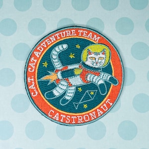 May include: Embroidered patch featuring a cat in a spacesuit, labeled "CAT ADVENTURE TEAM CATSTRONAUT." The patch has a teal background with an orange border. The cat is white with black stripes and a yellow helmet, with a rocket blasting from its side.