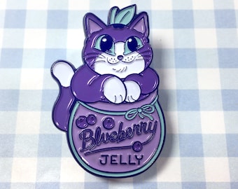 May include: Purple cat enamel pin with white accents, sitting inside a purple jar with the text "Blueberry Jelly" and three blueberries on the front.