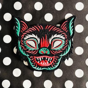 Könnte beinhalten: Schwarzer, roter und grüner gestickter Aufnäher mit einem Cartoon-Monster mit einem breiten Grinsen. Das Monster hat große Augen und eine spitze Nase.