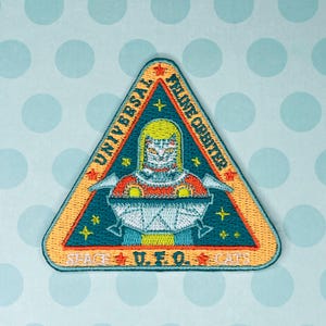 May include: Embroidered patch in a triangular shape with the text "UNIVERSAL FELINE ORBITER" and "SPACE U.F.O. CATS". The patch features a cat astronaut design in a teal, orange, yellow, and white color scheme. The background is light blue with white polka dots.