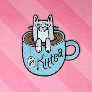 May include: Embroidered patch featuring a gray cat relaxing in a light blue teacup filled with brown liquid. The teacup has a handle and the word "Kittea" is written on the side. A tea bag with a leaf is also present.