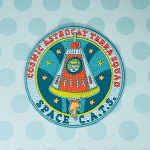 May include: Embroidered patch with the text "COSMIC ASTROCAT TERRA SQUAD SPACE C.A.T.S." featuring a rocket ship design in orange, white, and teal. The patch is circular with a teal background and yellow star accents.