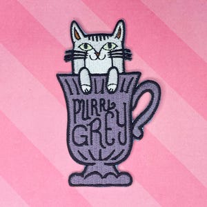May include: Embroidered patch featuring a gray cat peeking out of a purple teacup. The teacup has the words "Purr Grey" embroidered on it. The background is pink and white diagonal stripes.