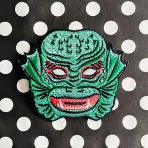 May include: Green embroidered patch of a creature from the black lagoon with red eyes and a toothy grin.