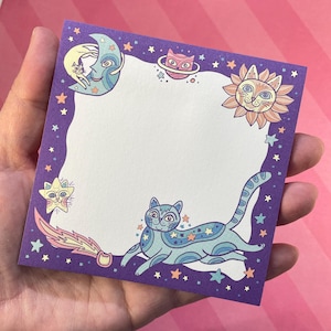 May include: A purple square notepad with a celestial cat design. The border features a cat-shaped sun, moon, stars, and planets. The central area is blank, ready for notes. The cat is blue and has star patterns.