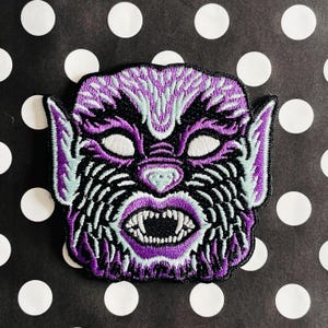 May include: Purple and blue embroidered patch of a monster face with sharp teeth and white eyes.