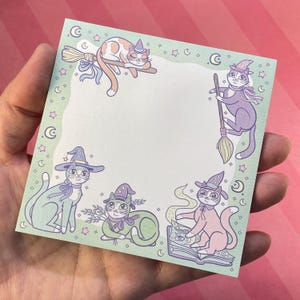 May include: A square notepad featuring whimsical illustrations of cats dressed as witches. The cats are depicted in various poses, including riding a broom, reading a spell book, and lounging. The notepad has a light green border with stars and crescent moons.