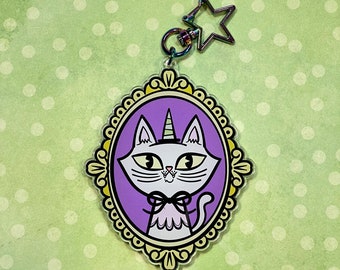 Peut inclure: Un porte-clés fantaisiste avec un chat-licorne dans un cadre ovale violet bordé de jaune. Le chat porte un nœud papillon et le porte-clés a un fermoir en forme d'étoile. Fond vert clair à pois blancs.