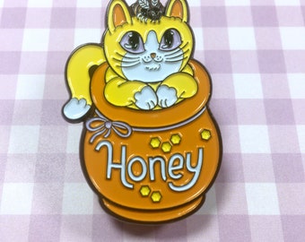 May include: A yellow cat with blue eyes is sitting inside a honey pot with the word "Honey" on the front. A small bee is perched on top of the cat's head.
