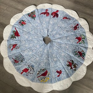 May include: A blue and white quilted Christmas tree skirt with a scalloped edge. The skirt features embroidered red cardinals, snowflakes, and the text "Merry & Bright".