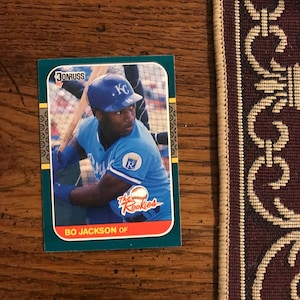 Può includere: Una carta di baseball di Bo Jackson del set Donruss del 1987. La carta presenta un'immagine a colori di Jackson con l'uniforme dei Kansas City Royals, con il testo "The Rookies" e "BO JACKSON OF". La carta ha un bordo verde.