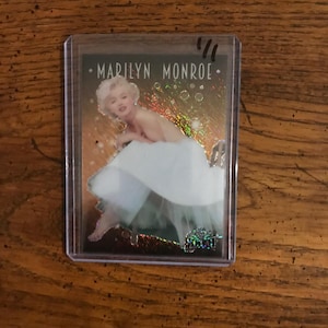 Op de afbeelding: Een verzamelkaart met een kleurenfoto van Marilyn Monroe in een witte jurk op een glinsterende achtergrond. De kaart heeft de tekst "Marilyn Monroe" erop gedrukt.