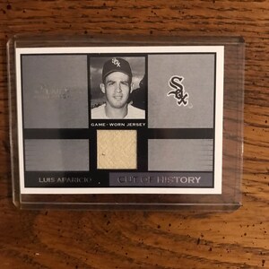Puede incluir: Una foto en blanco y negro de un jugador de béisbol que lleva una camiseta blanca con una "S" negra en la parte delantera. La foto está en una tarjeta con el texto "GAME-WORN JERSEY" y "CUT OF HISTORY". La tarjeta también tiene un pequeño trozo de la camiseta pegado a ella.