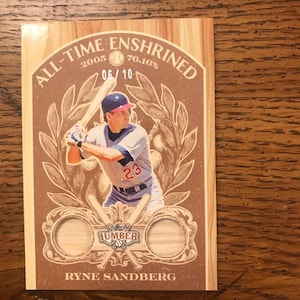 May include: A wooden baseball card with a baseball player in a blue and white uniform, holding a bat. The card has the text "ALL-TIME ENSHRINED" and "RYNE SANDBERG". The card also has two circular wooden inserts.