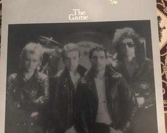 May include: A vinyl record album cover featuring a black and white photo of the band Queen. The album title "The Game" is printed in white on a gray background. The band members are wearing leather jackets and are looking at the camera.