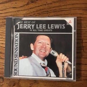 Puede incluir: Una caja de CD con "The Best of Jerry Lee Lewis: 16 All Time Greats." La portada muestra a un hombre sonriente con un micrófono. El texto "SOUND SENSATION" está impreso verticalmente a la izquierda. La caja es gris.