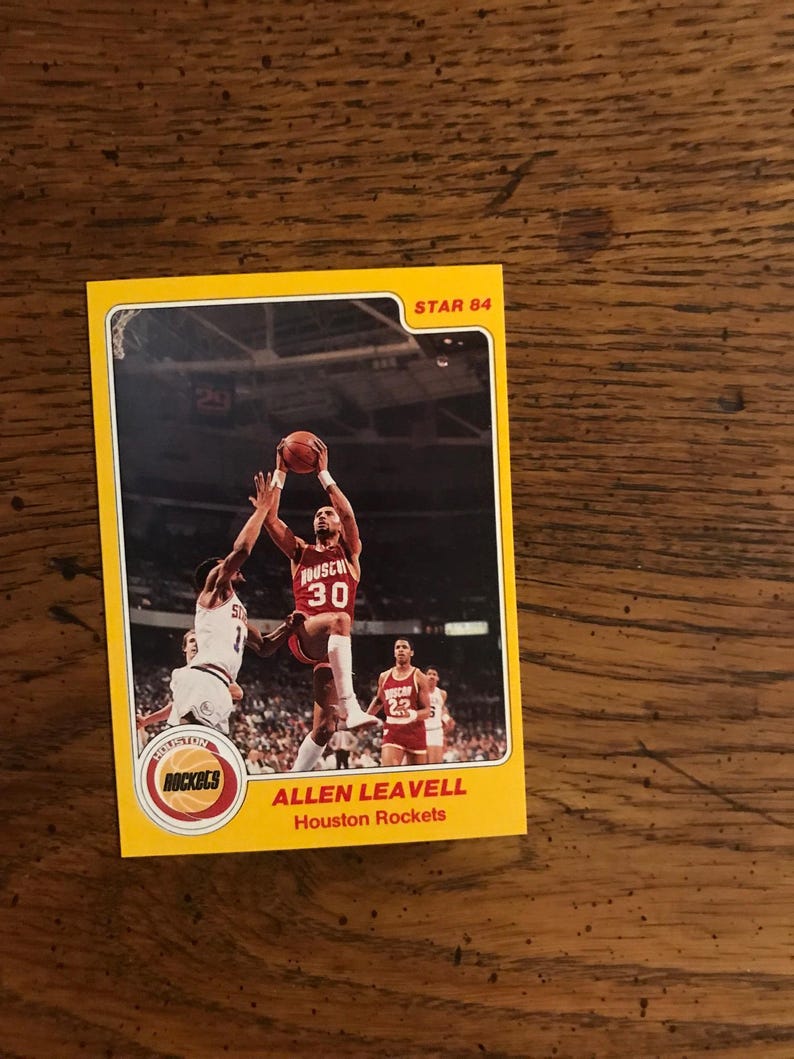 May include: A vintage Allen Leavell Houston Rockets basketball card. The card features a player in a red and white uniform, jumping to shoot a basketball. The card has a yellow border and the text "STAR 84".