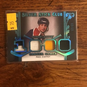 May include: A hockey trading card featuring Paul Coffey, with the text "Silver Stick Club" at the top. The card has a holographic design and includes the text "Superlative Career Games". The card is in a protective sleeve.
