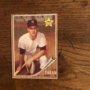Puede incluir: Una tarjeta de béisbol vintage con Tom Tresh, un jugador de los New York Yankees. La tarjeta es de 1962 y presenta una foto de Tresh con su uniforme, sosteniendo un bate. La tarjeta tiene una estrella amarilla con la palabra "Rookie" impresa en ella.