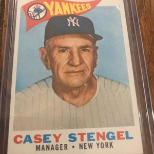 Puede incluir: Una tarjeta de béisbol vintage con un retrato de Casey Stengel usando una gorra azul oscuro de los New York Yankees. La tarjeta incluye un gráfico de banderín rojo con el texto YANKEES y el nombre CASEY STENGEL MANAGER NEW YORK impreso abajo.