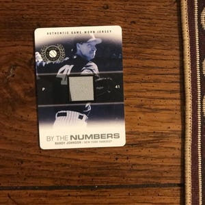 May include: A baseball card depicting Randy Johnson of the New York Yankees. The card shows Johnson in his uniform, with the number 41. The card's text includes "AUTHENTIC GAME-WORN JERSEY" and "BY THE NUMBERS".