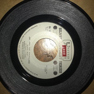 May include: A black and white 45 RPM vinyl record with a silver label. The label has the text "United Artists Records Inc. New York 19, N.Y. Made in U.S.A." and "Jay and the Americans "Cara Mia" (T. Trapani-L. Lange) Produced by: Garry Granahas Arranged by: Artie Goland". The label also has the text "United 1965 Silver Spotlight Series" and "UA 16".