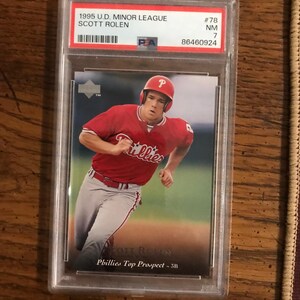 May include: A 1995 U.D. Minor League baseball card of Scott Rolen, graded and encased in a protective holder. The card features Rolen in a red Phillies uniform. Text on the card includes "Phillies Top Prospect - 3B" and grading information.