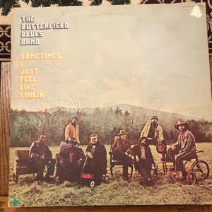 Puede incluir: Una portada de álbum de vinilo vintage con The Butterfield Blues Band. El arte de la portada muestra a los miembros de la banda sentados en un campo con una montaña al fondo. El título del álbum "Sometimes I Just Feel Like Smilin'" está impreso en blanco sobre un fondo azul claro.
