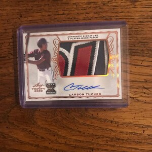 May include: A baseball trading card featuring Carson Tucker, with an authentic signature and player-worn patch. The card has a photo of Tucker in a baseball uniform and the text "Leaf Trinity 2020 Rookie". The patch has red, black, and white colours.