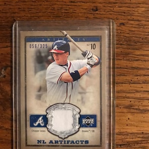Può includere: Una carta da baseball di Chipper Jones degli Atlanta Braves, in una custodia protettiva. La carta mostra il testo "ARTIFACTS" e "NL ARTIFACTS". Numerata 056/325, presenta il logo Upper Deck. Un oggetto da collezione.