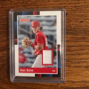 May include: A baseball card featuring Alec Bohm in a red uniform, with the Donruss logo in the upper left corner. The card is encased in a clear protective holder. The team abbreviation "PHI" is visible.