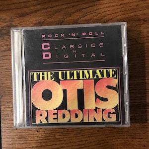 May include: A CD case with the album "The Ultimate Otis Redding." The cover is black with the text "Rock 'N' Roll Classics in Digital." The album title is in large orange and yellow letters with a pink border.