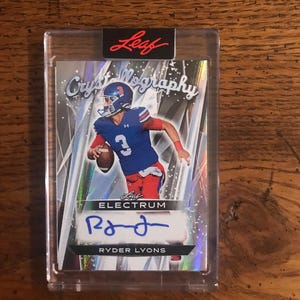May include: A football trading card encased in a clear protective sleeve. The card features a player in a blue and red uniform, holding a football. The card is signed and has the text "RYDER LYONS" at the bottom. The brand name "Leaf" is at the top.