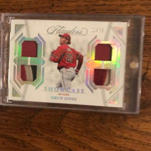 Puede incluir: Una tarjeta de béisbol con Druw Jones con uniforme rojo de Arizona. La tarjeta tiene dos parches y la palabra "Flawless" en la parte superior. La tarjeta está dentro de un protector transparente.