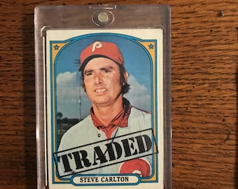 May include: A vintage baseball card of Steve Carlton, in a protective case. The card features a portrait of Carlton in a Phillies uniform, with the word "TRADED" stamped across the front. The card has a blue and yellow border.