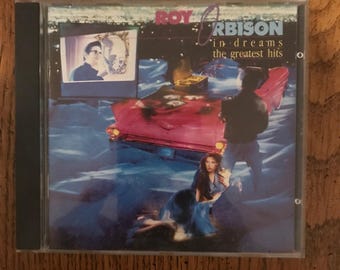 May include: A CD case featuring a pink car and a woman in a blue dress on the cover. The album title is "Roy Orbison In Dreams The Greatest Hits".