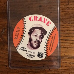 May include: A white baseball card with a red border featuring Willie Stargell, an outfielder for the Pittsburgh Pirates. The card has the text "CRANE" in red letters and "MLB" in red and white letters. The card also includes Stargell's height, weight, and date of birth.