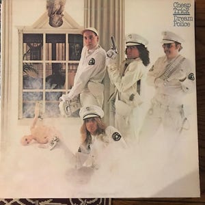 May include: Album cover for Cheap Trick's "Dream Police" featuring the band members in white uniforms and hats, posing with a doll and a gun. The album title is in the upper right corner.