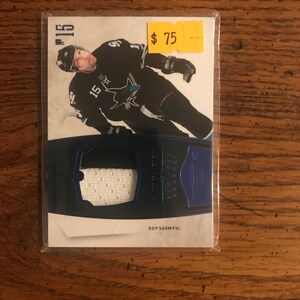 May include: A hockey trading card featuring a player in a black and teal uniform with the number 15. The card has a patch insert and the word "NAMEPLATE" printed at the bottom. The card is encased in a protective sleeve.